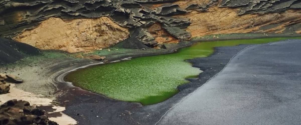A short distance along the southwest coastline from the exciting Los Hervideros is a colourful natural example of the power of Mother Nature. El Golfo is a half-submerged cone of a volcano, which over time has been eroded by the sea, leaving behind only the striated wall of the crater displaying a myriad of red and russet colours.
At the foot of the crater wall is Lago Verde, a half-moon-shaped striking green lagoon filled with volcanic minerals and micro-organisms that are believed to be unique in this lake.
A beach of black volcanic pebbles, where you can look for specimens of the semi-precious olivine, superficially separates the sharply contrasting lagoon from the blue waters of the Atlantic Ocean, but in reality they are connected through underground passageways.
Coming from the south, the road leads you to a small car park, from where you can approach El Golfo by foot, following signs for a short walk around the headland.
A number of sheltered bays with black beaches link the lagoon with the tranquil El Golfo village, which attracts geologists and jewellery makers because of the olivine found in this area.
In addition, this small and peaceful community boasts several excellent seafood restaurants – great places to enjoy a tasty meal while enjoying a beautiful sunset and magnificent views.