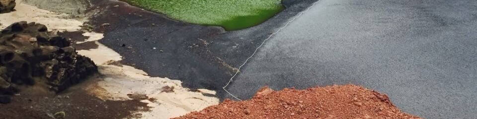 A short distance along the southwest coastline from the exciting Los Hervideros is a colourful natural example of the power of Mother Nature. El Golfo is a half-submerged cone of a volcano, which over time has been eroded by the sea, leaving behind only the striated wall of the crater displaying a myriad of red and russet colours.
At the foot of the crater wall is Lago Verde, a half-moon-shaped striking green lagoon filled with volcanic minerals and micro-organisms that are believed to be unique in this lake.
A beach of black volcanic pebbles, where you can look for specimens of the semi-precious olivine, superficially separates the sharply contrasting lagoon from the blue waters of the Atlantic Ocean, but in reality they are connected through underground passageways.
Coming from the south, the road leads you to a small car park, from where you can approach El Golfo by foot, following signs for a short walk around the headland.
A number of sheltered bays with black beaches link the lagoon with the tranquil El Golfo village, which attracts geologists and jewellery makers because of the olivine found in this area.
In addition, this small and peaceful community boasts several excellent seafood restaurants – great places to enjoy a tasty meal while enjoying a beautiful sunset and magnificent views.