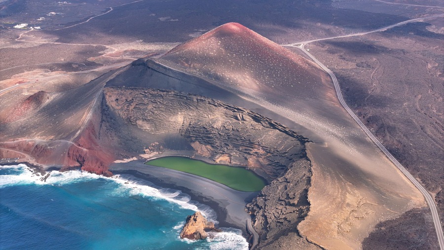 An aerial view of a volcanic crater with a green lake, surrounded by rugged terrain and a coastal road.