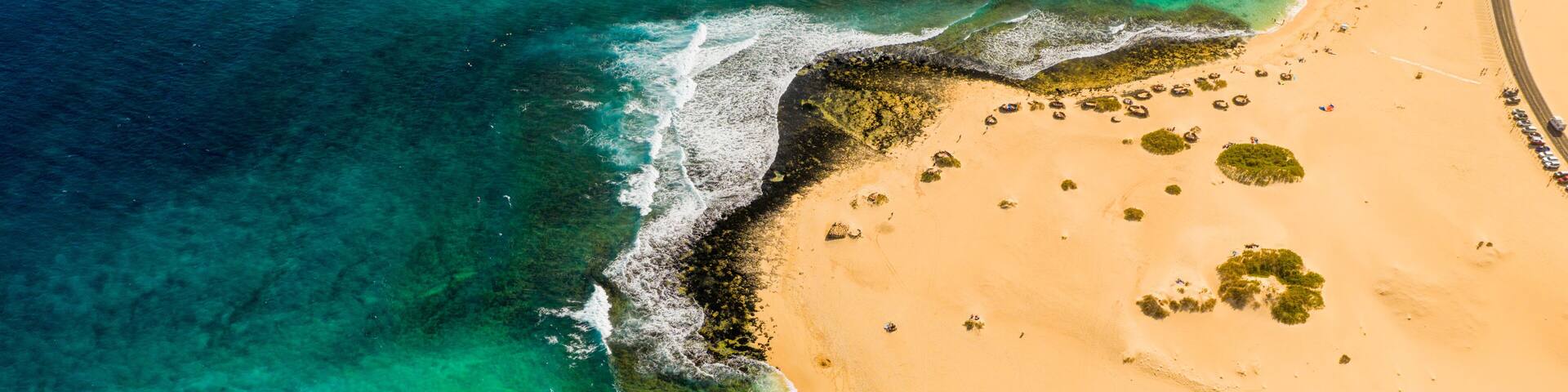 An aerial view of a beach with clear turquoise waters, a sandy shore, and scattered vegetation.