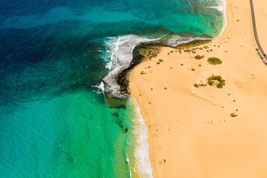 An aerial view of a beach with clear turquoise waters, a sandy shore, and scattered vegetation.