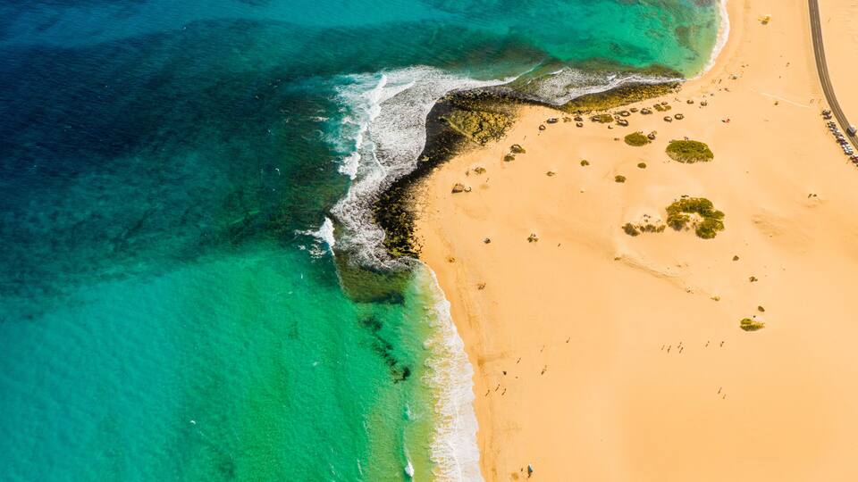 An aerial view of a beach with clear turquoise waters, a sandy shore, and scattered vegetation.