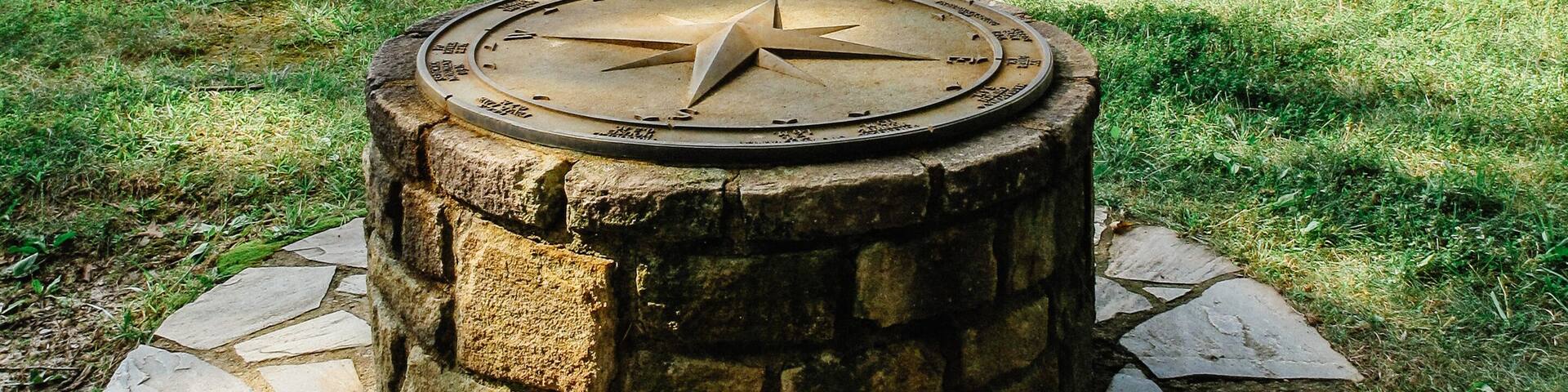 A stone compass on a circular stone platform surrounded by grass and trees.