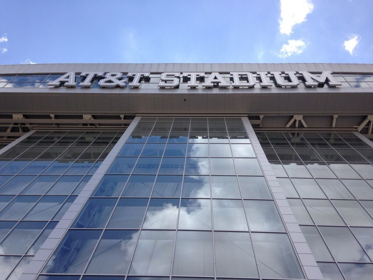 The AT&T Stadium with a clear sky and clouds reflected in the glass facade.