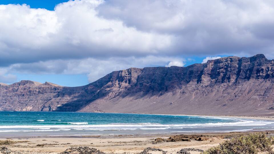 A coastal landscape with a rocky beach, a flag on a pole, and a mountain range in the background.