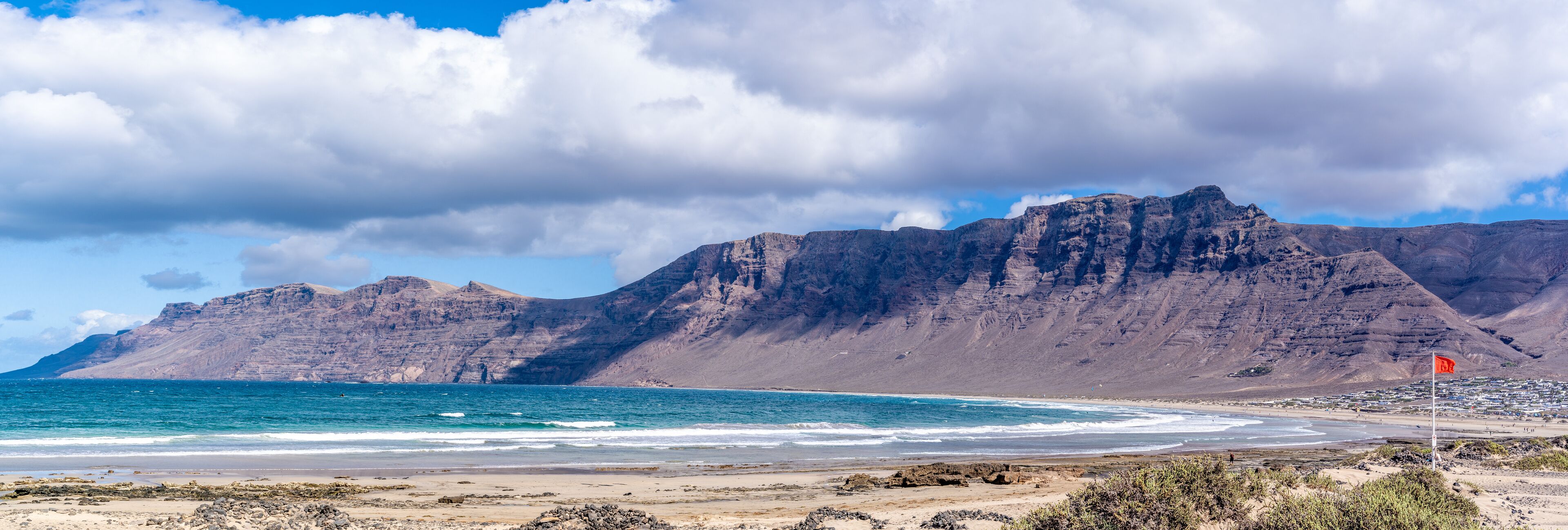A coastal landscape with a rocky beach, a flag on a pole, and a mountain range in the background.