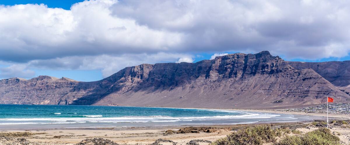 A coastal landscape with a rocky beach, a flag on a pole, and a mountain range in the background.