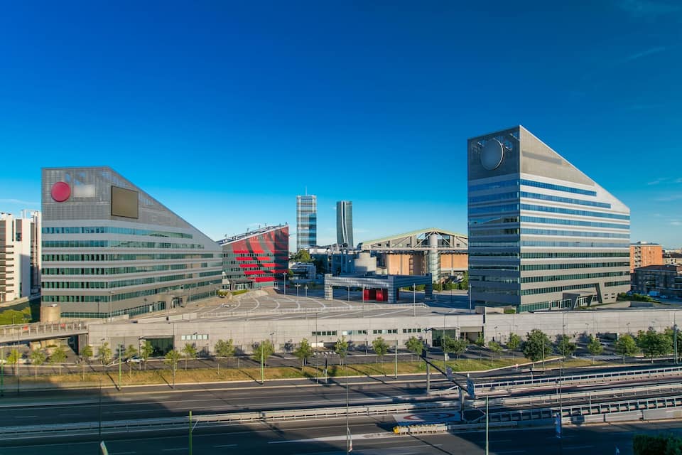 Paisagem urbana moderna com arquitetura marcante, incluindo um edifício com um detalhe circular vermelho e outro com uma torre de relógio.