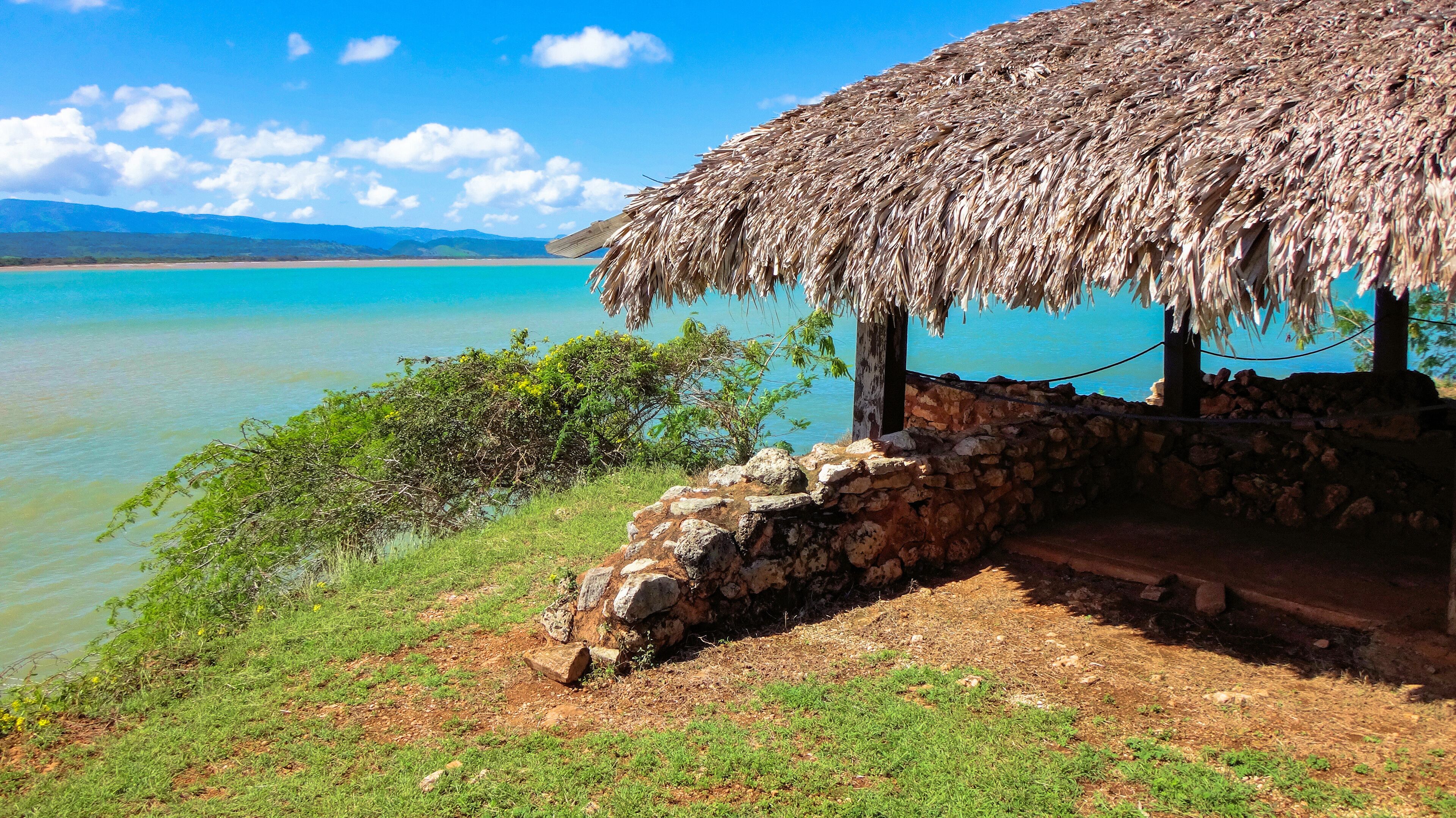 A thatched-roof hut by a turquoise lake with a stone wall and greenery.