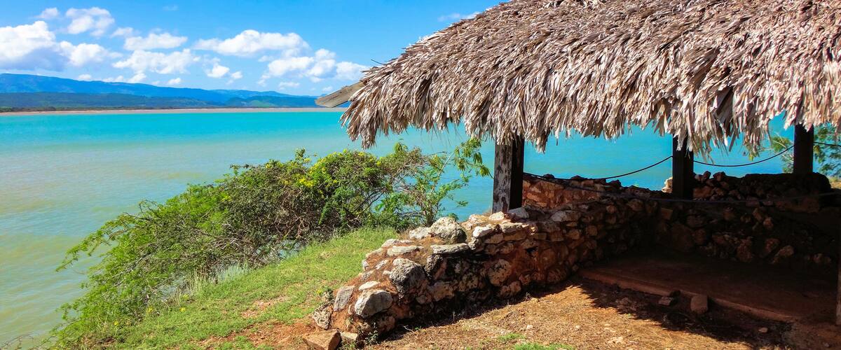 A thatched-roof hut by a turquoise lake with a stone wall and greenery.
