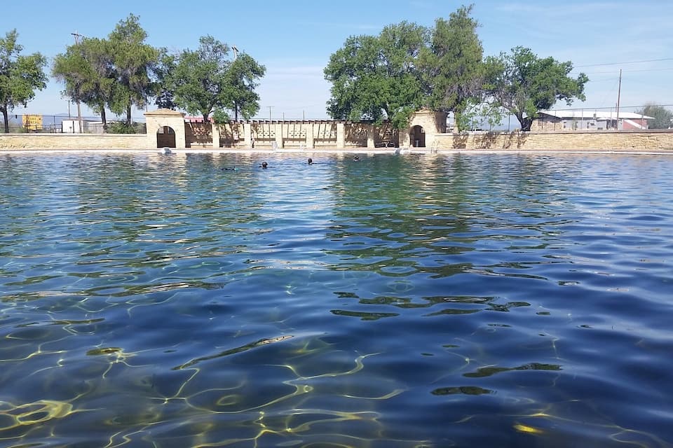 Um lago calmo com águas cristalinas, rodeado por árvores e um muro de pedra com arcos.