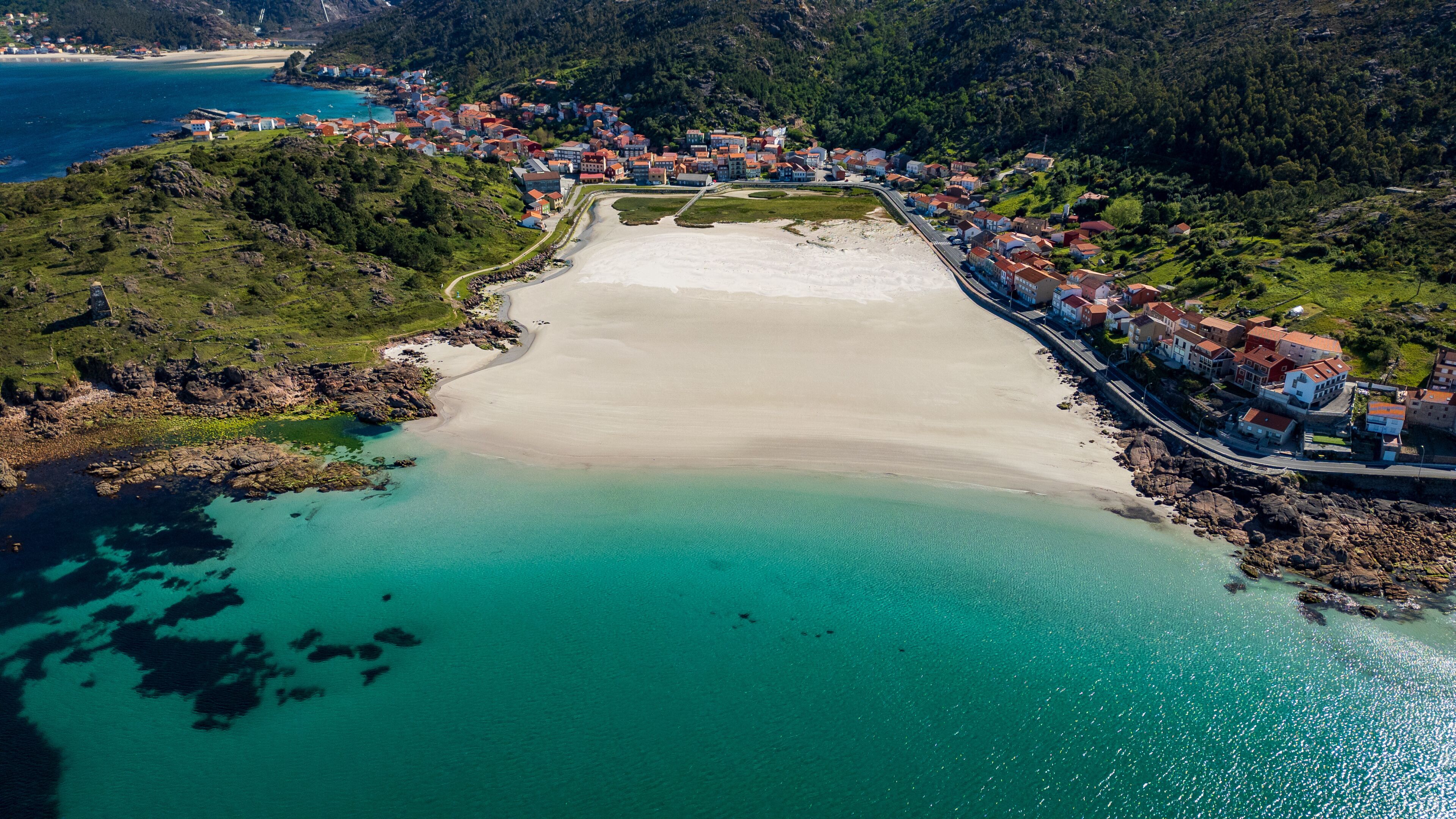 A coastal town with a sandy beach, clear turquoise waters, and red-roofed buildings.