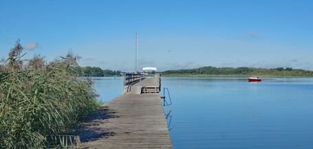 Eine Holzstege, der sich in ruhiges Wasser erstreckt, ein rotes Boot und eine ferne Hügelkette mit einer hohen Struktur.