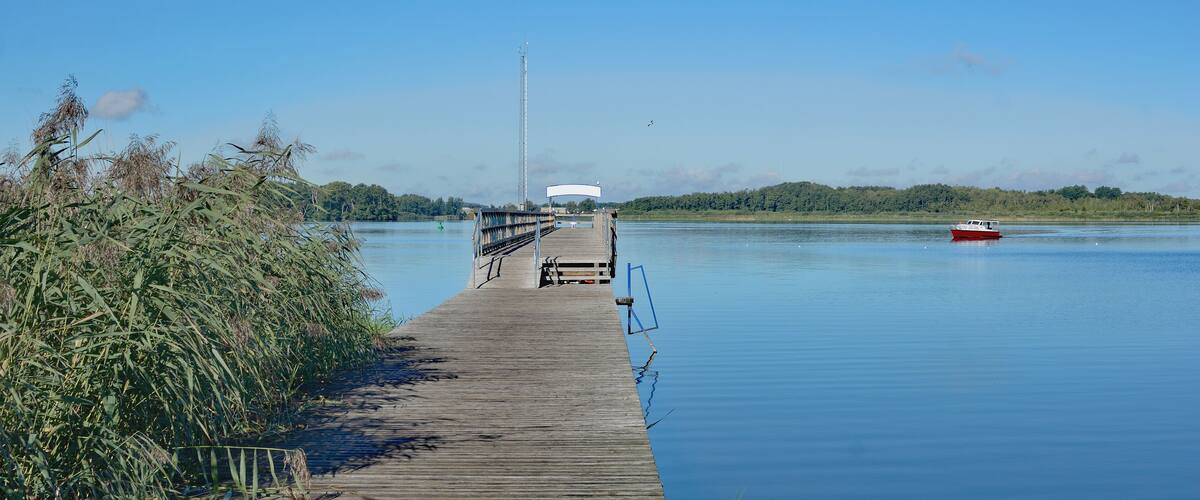 A wooden pier extending into calm water, a red boat, and a distant hill with a tall structure.