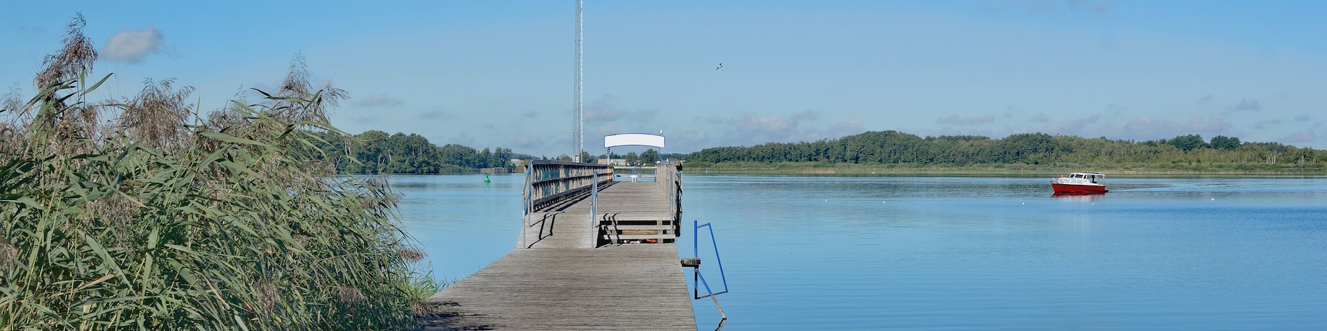 A wooden pier extending into calm water, a red boat, and a distant hill with a tall structure.