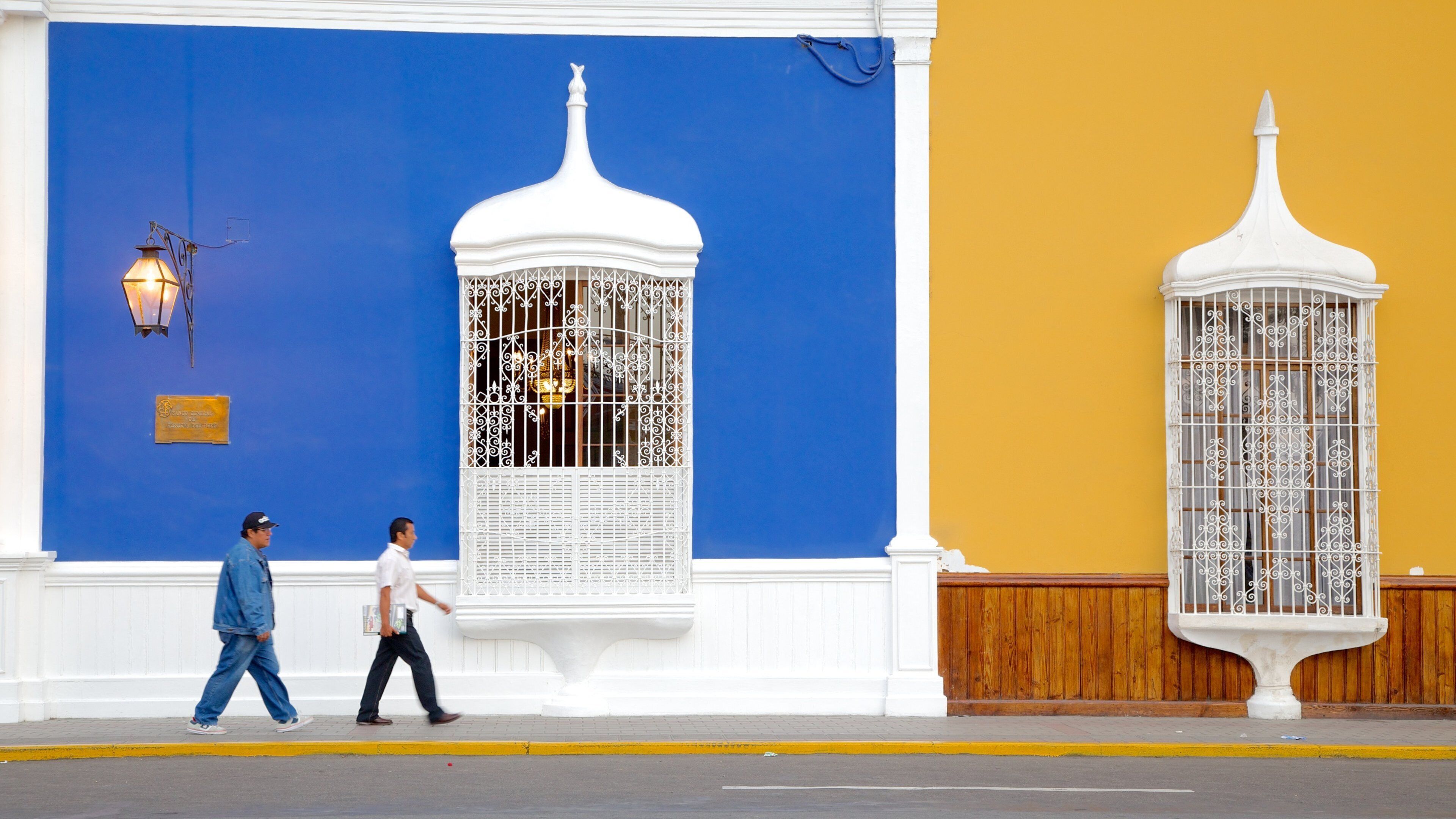 A building facade with blue and yellow walls, white decorative elements, and a street with pedestrians.