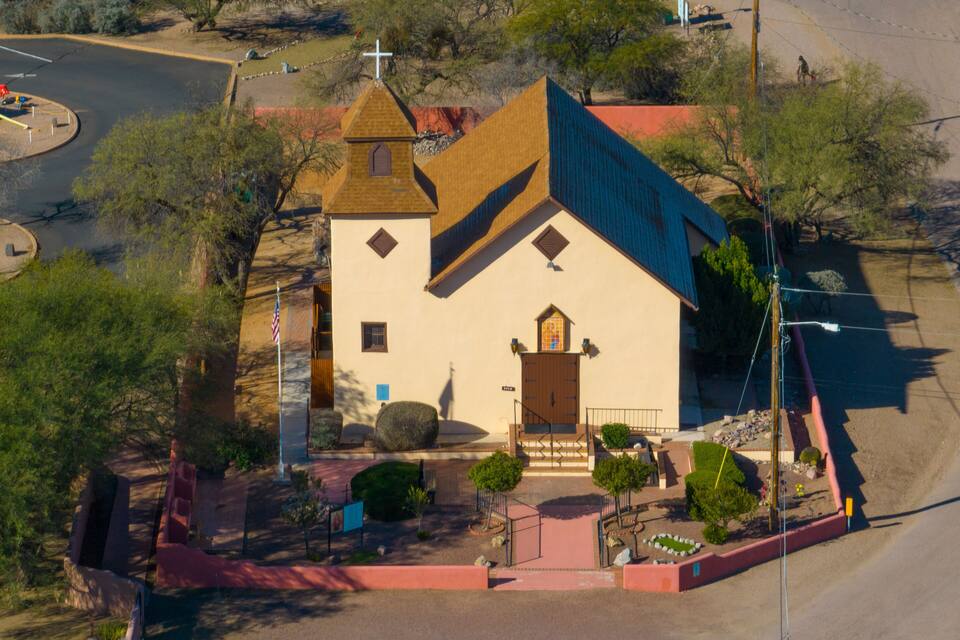 A small church with a cross on top, surrounded by trees and a paved area.