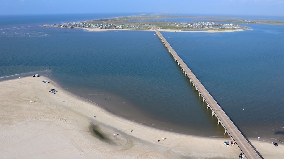 A coastal area with a long pier extending into the water, a sandy beach, and a small island with buildings in the distance.