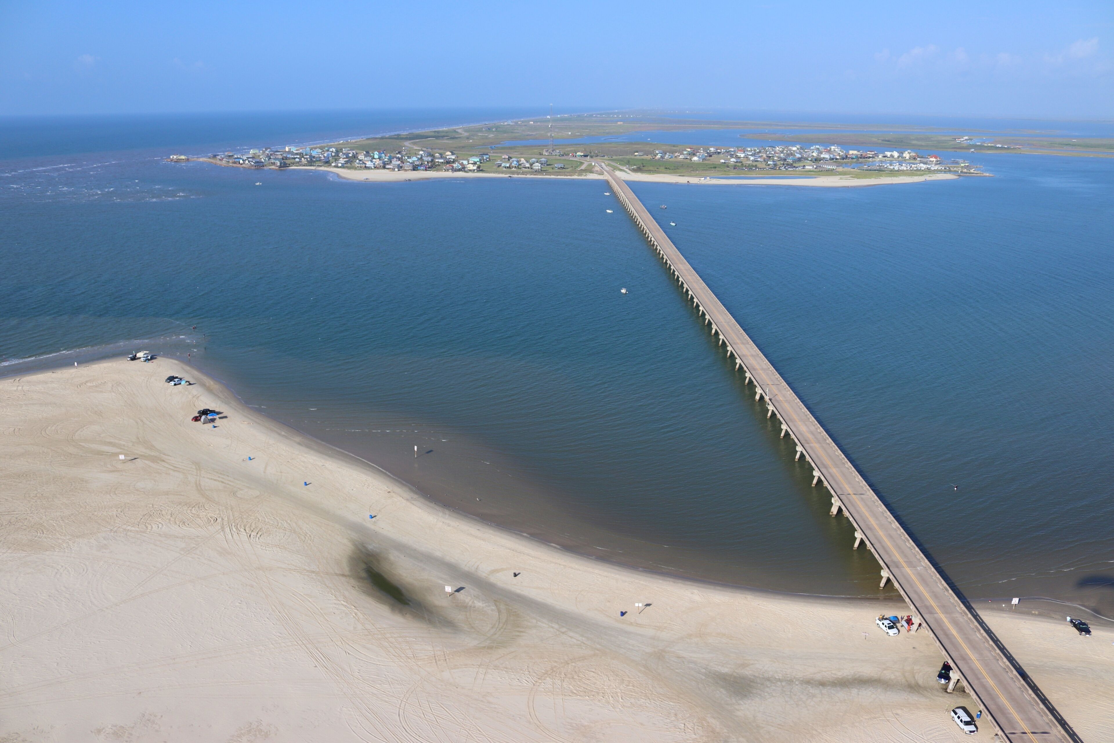 A coastal area with a long pier extending into the water, a sandy beach, and a small island with buildings in the distance.