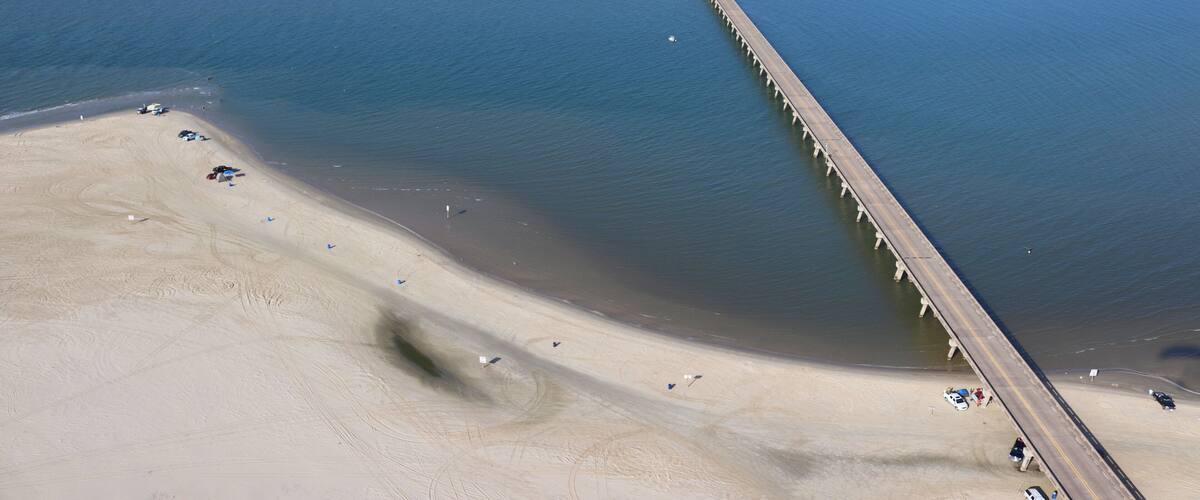 A coastal area with a long pier extending into the water, a sandy beach, and a small island with buildings in the distance.