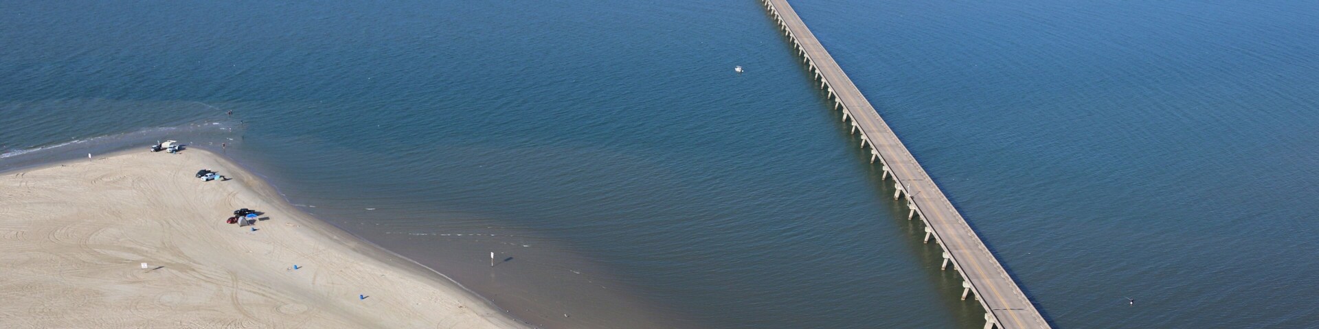 A coastal area with a long pier extending into the water, a sandy beach, and a small island with buildings in the distance.