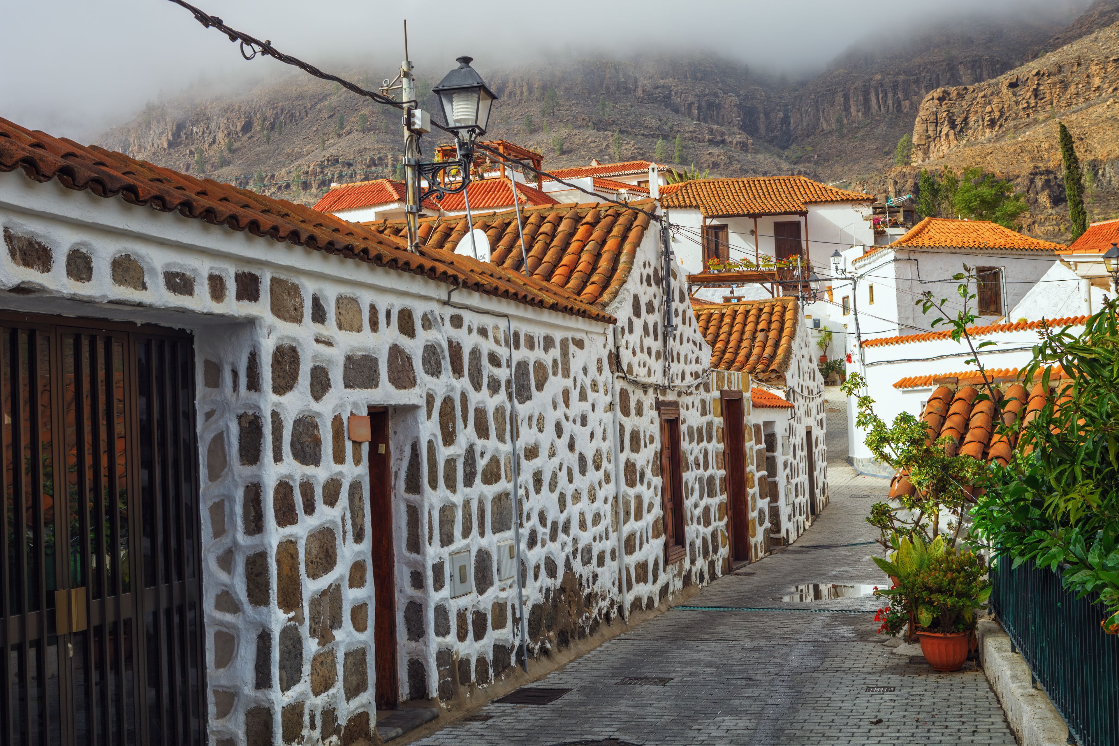A narrow cobblestone street lined with white buildings with red-tiled roofs, set against a mountainous backdrop.