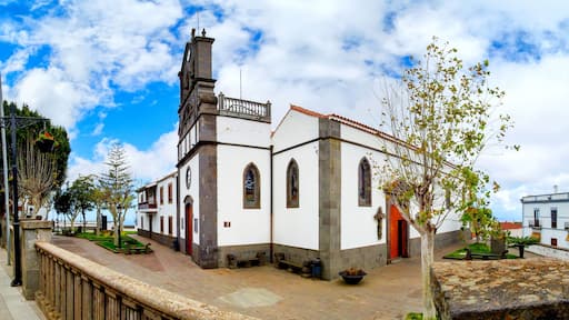 Igreja branca com torre de sino e porta vermelha, cercada por árvores e um caminho pavimentado.