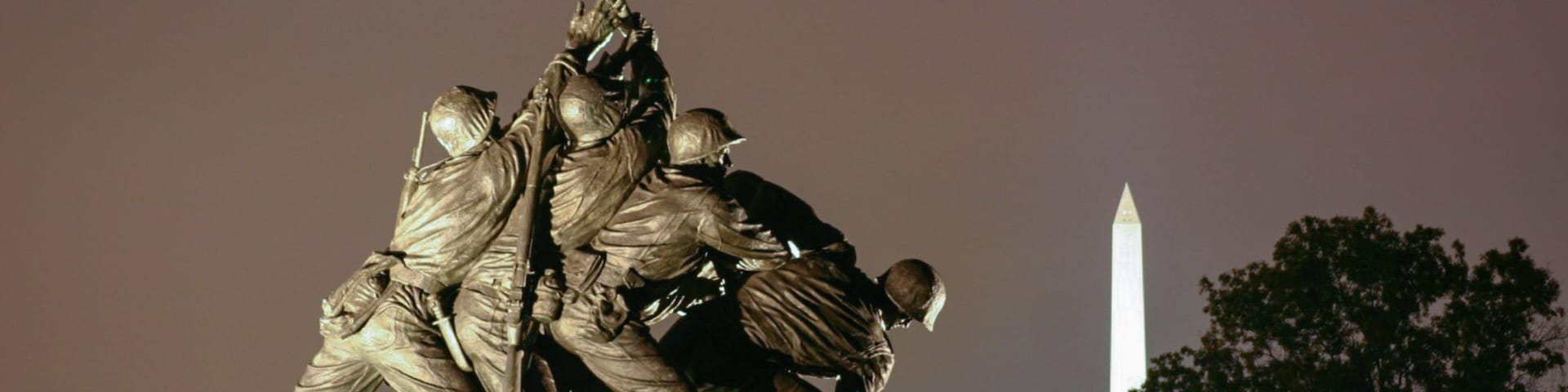 Estátua noturna de soldados erguendo uma bandeira, com o Monumento a Washington e o Capitólio ao fundo.