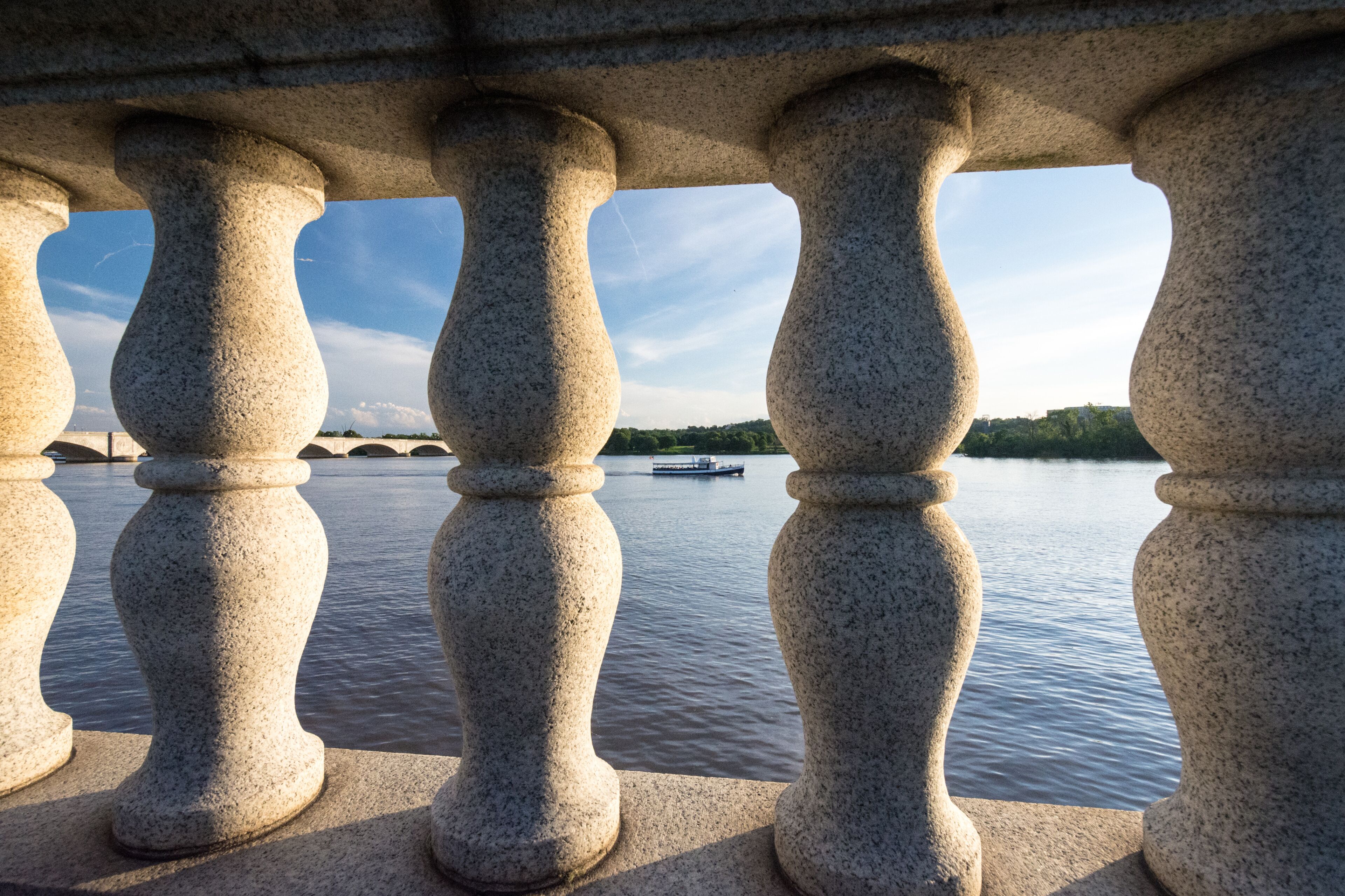 A stone balustrade overlooking a calm river with a bridge in the distance.