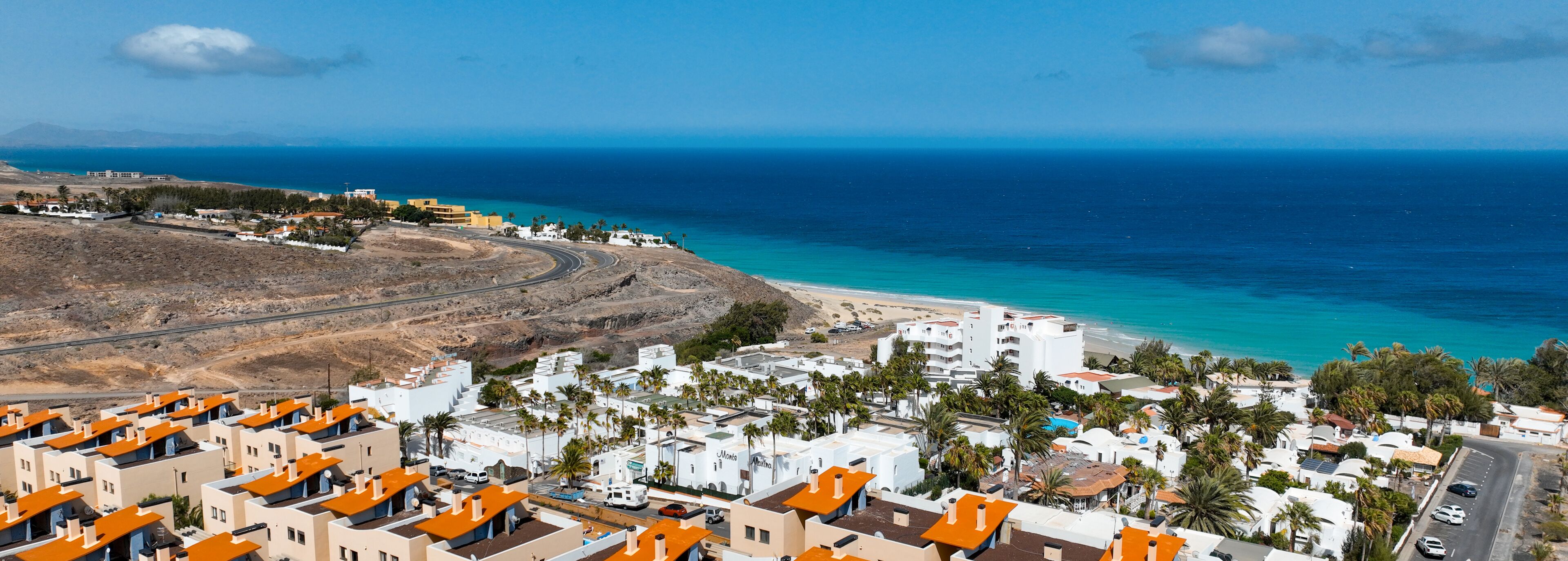 A coastal town with white buildings and orange roofs, a winding road, and a beach with clear blue waters.