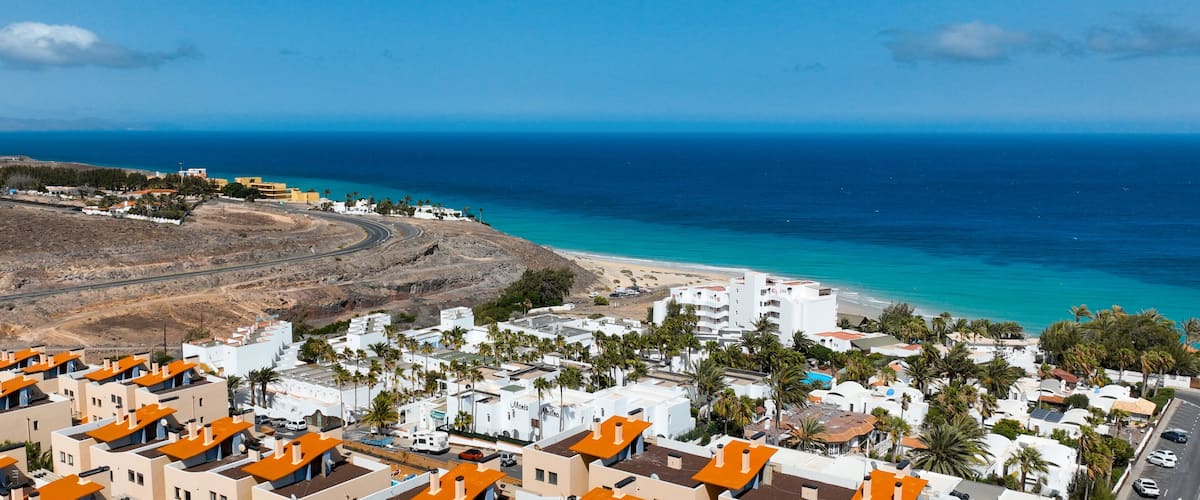 A coastal town with white buildings and orange roofs, a winding road, and a beach with clear blue waters.