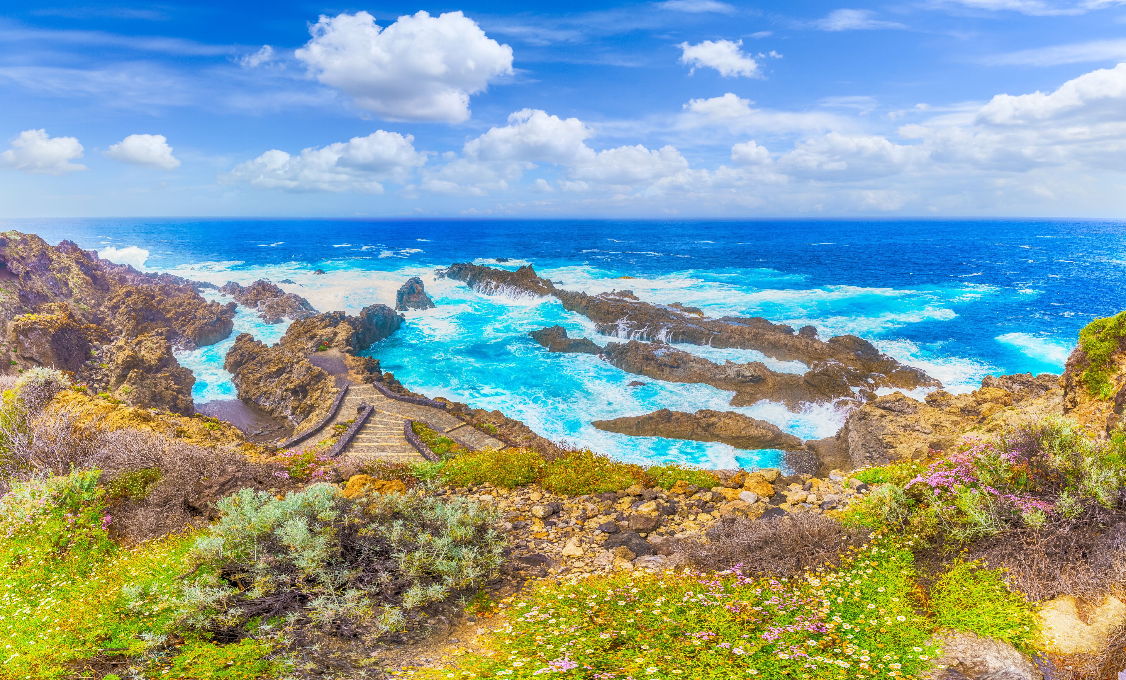 A coastal landscape with rocky cliffs, a sea view, and a stone staircase leading down to the water.
