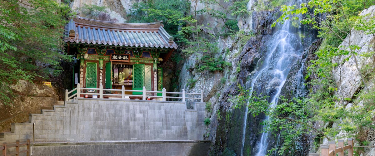 A traditional temple with a tiled roof, situated at the base of a waterfall in a rocky gorge.