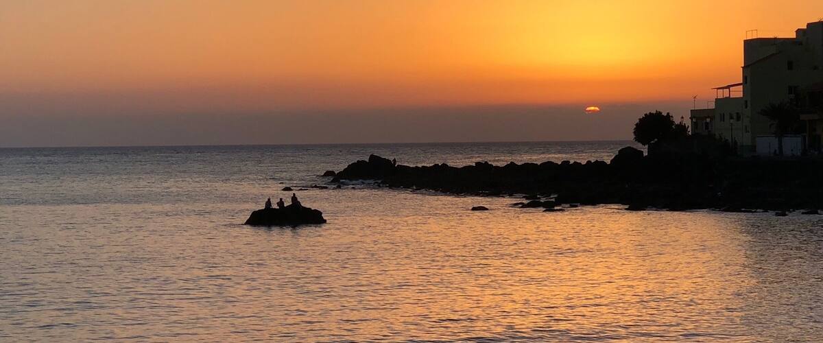 A sunset over a calm sea with rocky outcrops and buildings on the right.