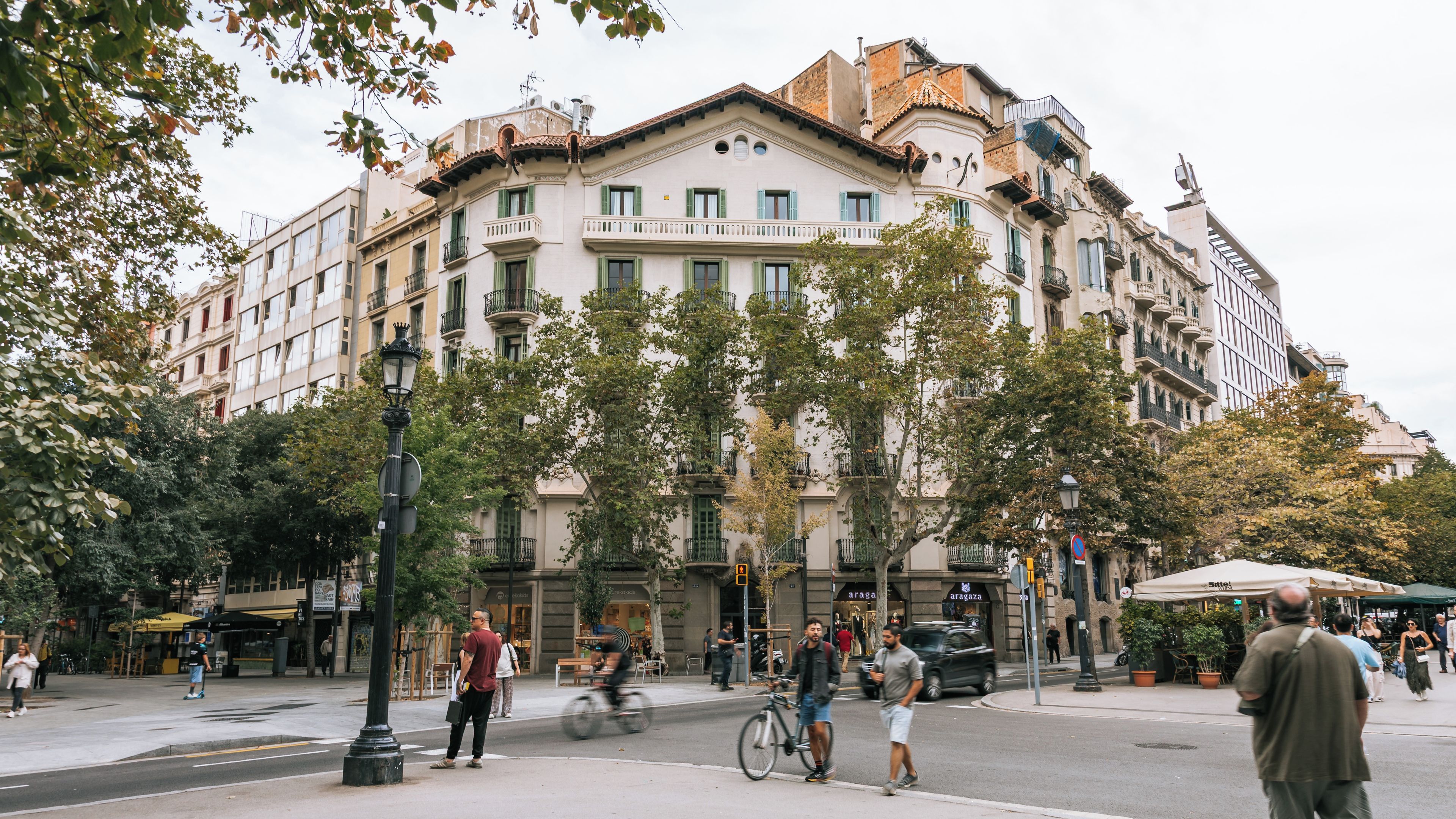 Rua com pedestres, ciclistas e carros. Árvores margeiam a calçada, e um edifício grande com várias sacadas e janelas.