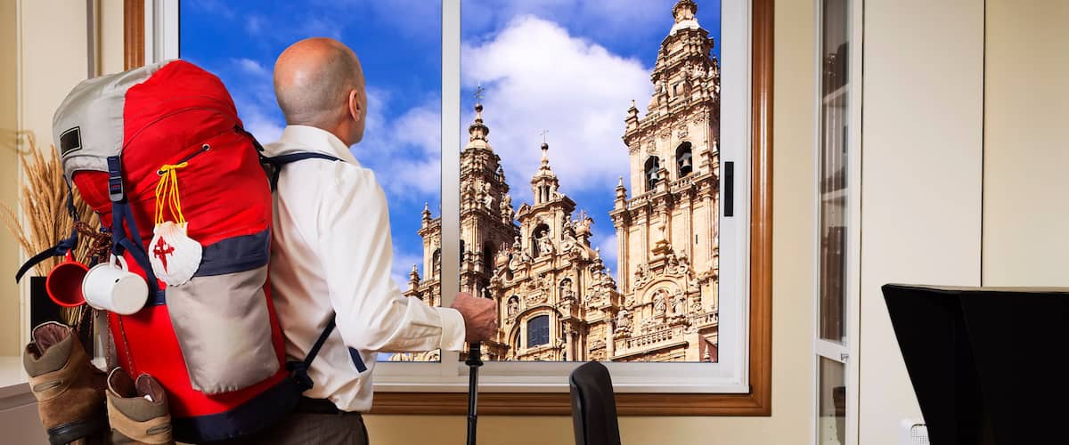man in office with backpack and trekking boots looking trought the window dreaming of trekking towards Santiago of Compostela