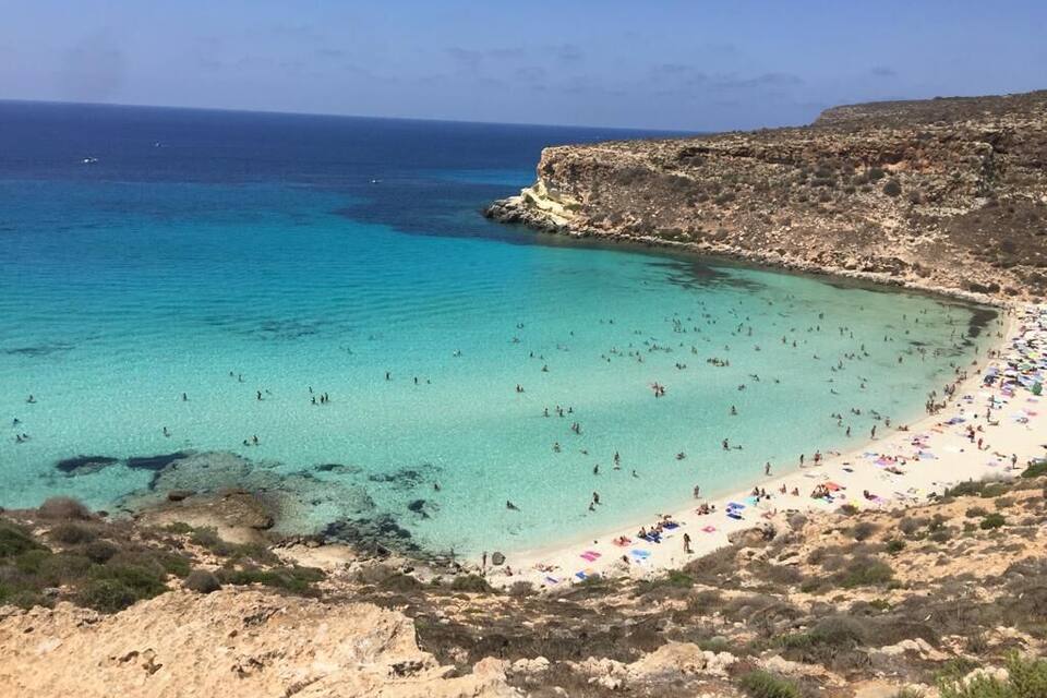 Una playa costera con aguas turquesas cristalinas, acantilados rocosos y gente tomando el sol.