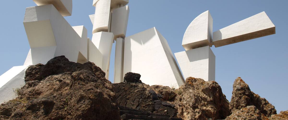 A white abstract sculpture with geometric shapes, set against a clear blue sky, on a rocky terrain.