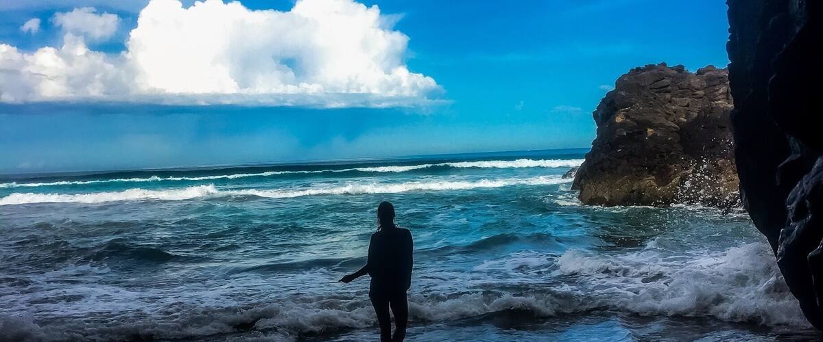 A person walking in the ocean near a rocky cliff under a partly cloudy sky.