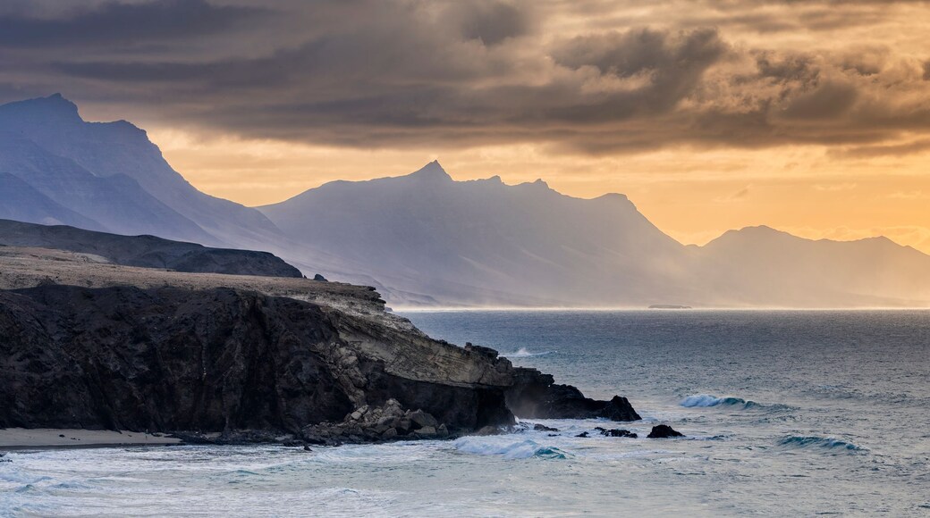 A coastal landscape with rugged mountains, a rocky shoreline, and a calm sea under a dramatic sky.