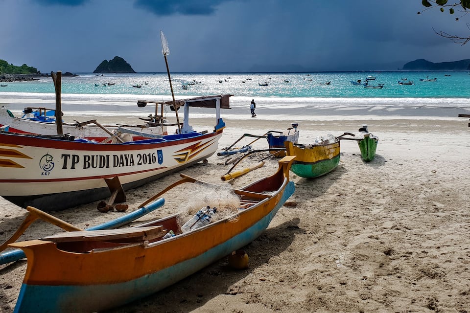 Une plage où se trouvent plusieurs bateaux, dont un bateau rouge et blanc sur lequel on peut lire « TP BUDI DAYA 2016 », ainsi qu’un bateau bleu et jaune.