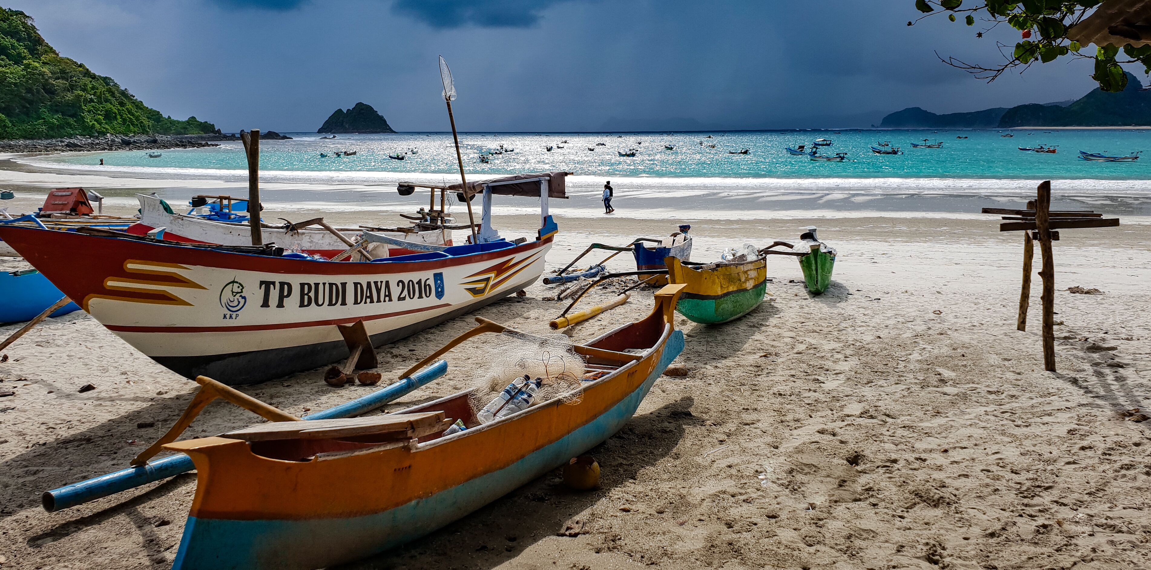 A beach with several boats, including a red and white boat with 'TP BUDI DAYA 2016' written on it, and a blue and yellow boat.