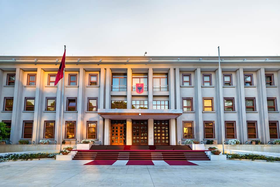 Un edificio grande y moderno con una bandera roja en primer plano, rodeado de follaje.