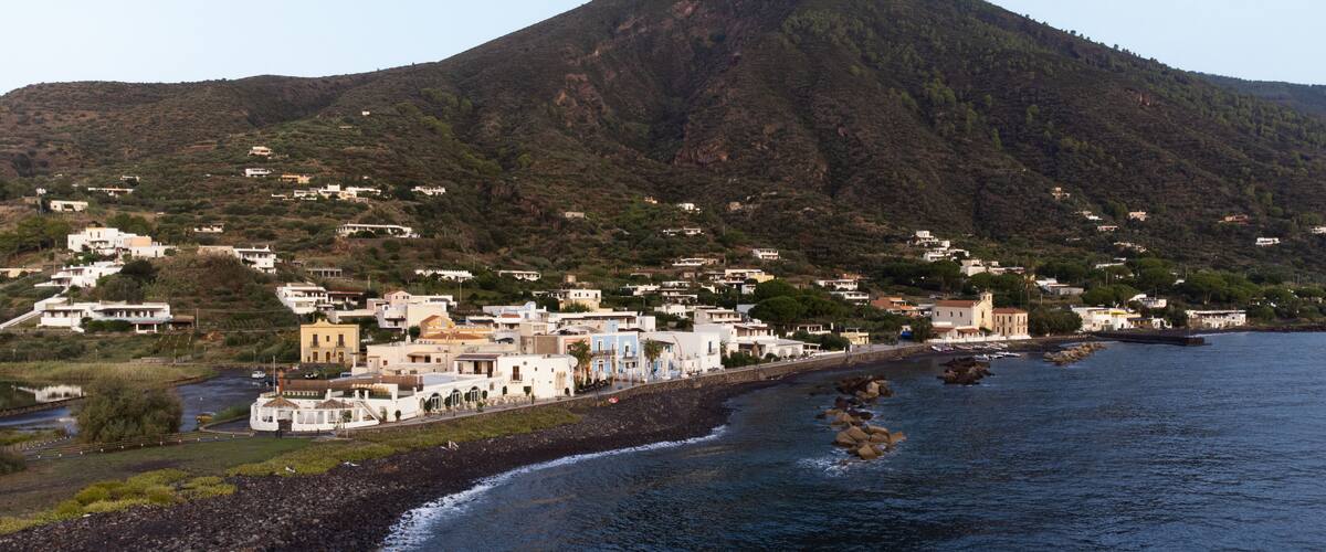 A coastal town with buildings along the shoreline, a mountain in the background, and rocky outcrops in the water.