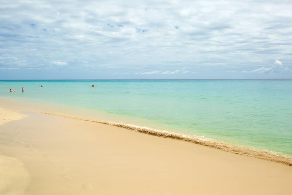 A sandy beach with clear turquoise water and a few people walking along the shore.