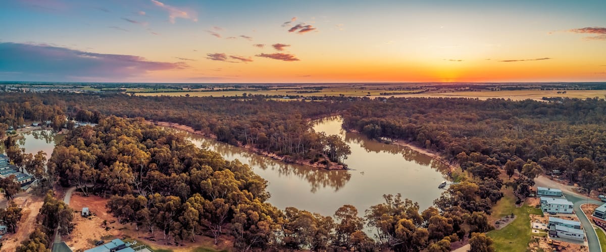 A river winding through a forested area with houses and a sunset in the background.