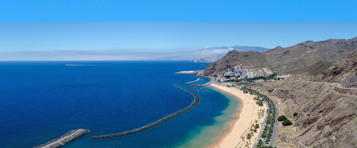 A coastal landscape with a beach, a curving road, and a rocky mountain range.