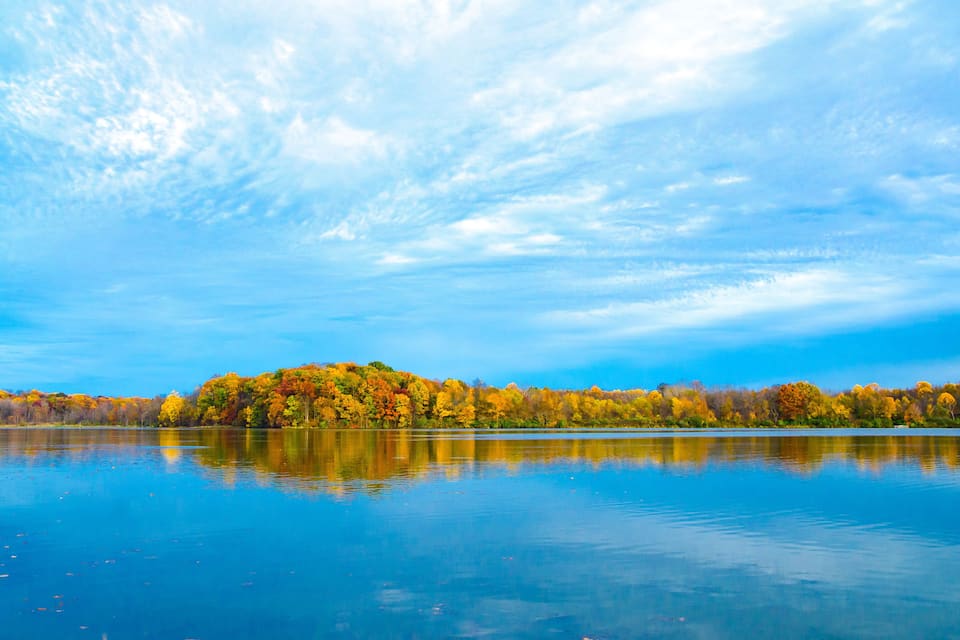 Een rustig meer met herfstbomen, wier spiegelbeeld in het water zichtbaar is.