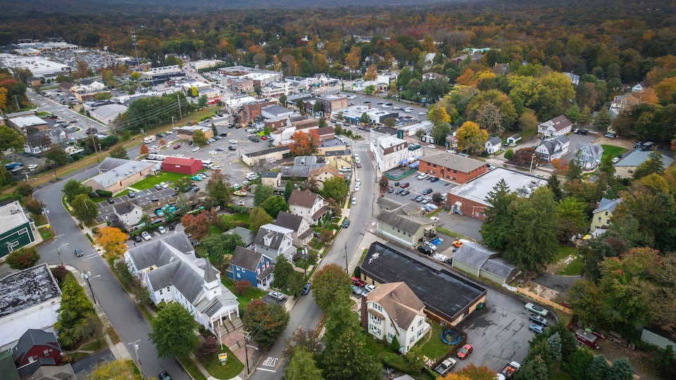 An aerial view of a town with a mix of residential and commercial buildings, surrounded by autumn-colored trees.