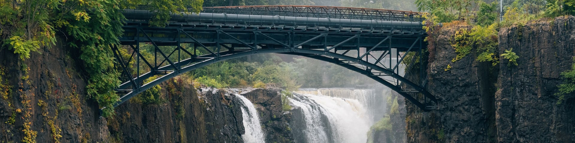 A bridge over a waterfall with surrounding rocky cliffs and greenery.